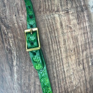 May include: Close-up of a green leather strap with leaf-shaped cutouts and a brass buckle. The strap is dyed a vibrant green color and is set against a wooden background. The buckle is rectangular and has a shiny, golden finish.