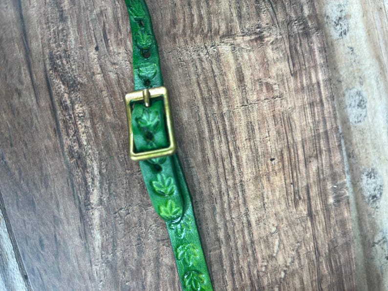May include: Close-up of a green leather strap with leaf-shaped cutouts and a brass buckle. The strap is dyed a vibrant green color and is set against a wooden background. The buckle is rectangular and has a shiny, golden finish.