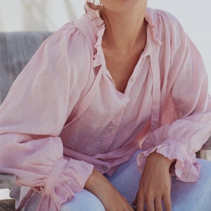 May include: A woman wearing a light pink, ruffled blouse with long sleeves and a button-up front.
