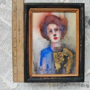 May include: A watercolor painting of a person with red hair and blue eyes wearing a blue shirt and a gold-colored scarf. The painting is framed in a black frame with a gold mat.