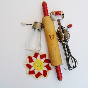 May include: A collection of vintage kitchen tools, including a wooden rolling pin with red handles, a hand-cranked egg beater, and a glass salt shaker. A red, white, and yellow crocheted trivet is also included. The items are arranged on a white surface.