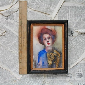 May include: A watercolor painting of a person with red hair wearing a blue and gold outfit. The painting is framed in black with a gold inner frame. The artist's signature is visible in the bottom left corner.
