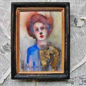 May include: A watercolor painting of a woman with red hair and blue eyes. She is wearing a blue shirt and a gold necklace. The painting is framed in a black frame.