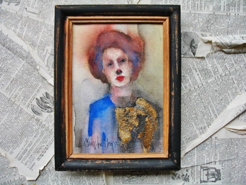 May include: A watercolor painting of a woman with red hair and blue eyes. She is wearing a blue shirt and a gold necklace. The painting is framed in a black frame.