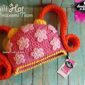 May include: A handmade crochet hat in pink with light pink flower appliques, yellow trim, and orange accents. The hat has a unique design with two orange loops extending from the sides. Text on the hat reads "Milli Hat Umizoomi Team".