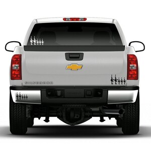 May include: White pickup truck with black and white rifle decals on the rear window and bumper. The truck has a silver body and a gold Chevrolet emblem. The word "SILVERADO" is printed across the tailgate.