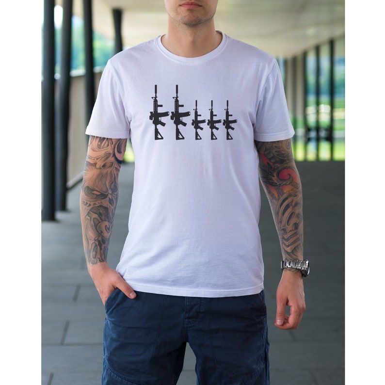 May include: White t-shirt featuring a black graphic of five rifles in a row. The t-shirt is short-sleeved and appears to be made of cotton. The person wearing the shirt has tattoos on both arms and a watch on their left wrist.