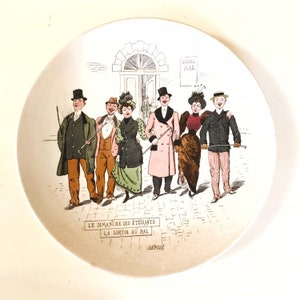 May include: A vintage ceramic plate featuring a colourful illustration of a group of people in 19th-century attire walking down a street. The text on the plate reads "Le Dimanche des Étudiants La Sortie du Bal."
