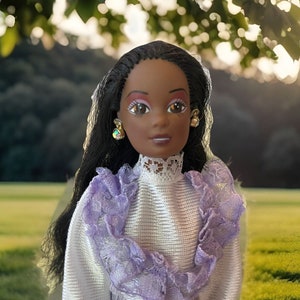 Puede incluir: Una muñeca con el pelo largo y negro, que lleva un vestido blanco con ribetes de encaje morado. Tiene los ojos marrones con sombra de ojos rosa y lleva pendientes de oro.