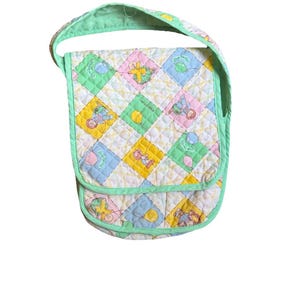 Puede incluir: Un bolso bandolera acolchado con correa y ribete verde claro. El bolso presenta un diseño de patchwork con cuadrados pastel de color amarillo, rosa, azul y verde, cada uno con un gráfico con temática de bebé. El bolso está diseñado para transportar objetos.