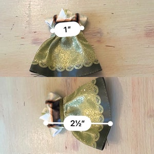 May include: A green and gold glittery dress with a brown trim and white accents. The dress measures 1 inch wide and 2 1/2 inches long.