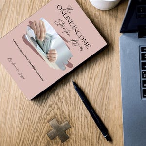 May include: A book titled "The Online Income Starter Kit" with the subtitle "7 Proven Paths to Your First $500 From Home" is displayed on a wooden surface. A laptop, pen, and decorative wooden cross are also visible.