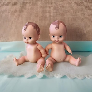 Peut inclure: Deux poupées de bébé en plastique vintage avec des cheveux et des yeux marrons peints. Les poupées sont assises sur une surface en tissu bleu clair et blanc.