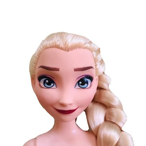 K&ouml;nnte beinhalten: Eine Puppe mit blonden Haaren, die zu einem Zopf geflochten sind, blauen Augen und einem gemalten L&auml;cheln. Die Puppe hat einen hellrosa Teint und tr&auml;gt keine Kleidung.