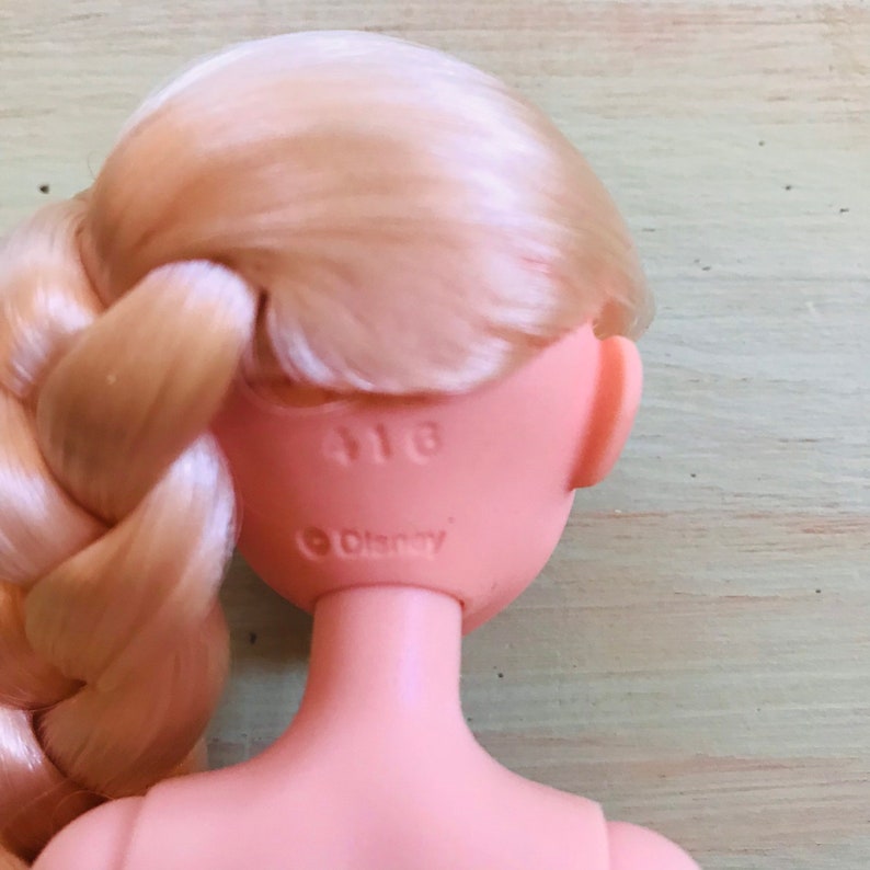 K&ouml;nnte beinhalten: Die R&uuml;ckseite des Kopfes einer blonden Puppe mit einem Zopf. Die Puppe hat einen rosa Hals und auf der R&uuml;ckseite ihres Kopfes befindet sich ein Aufkleber mit der Aufschrift "Disney" und "418".