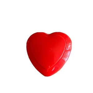 Può includere: Oggetto rosso a forma di cuore con finitura lucida. Il design presenta una forma a cuore più piccola all'interno di una più grande. La superficie riflette la luce, suggerendo una consistenza liscia, forse in plastica o laccata.