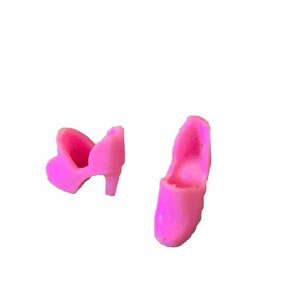 Puede incluir: Dos zapatos de muñeca de plástico rosa. Un zapato tiene tacón y el otro es plano.