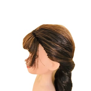 May include: A doll's head with long brown hair styled in a braid. The hair has highlights of a lighter brown colour.