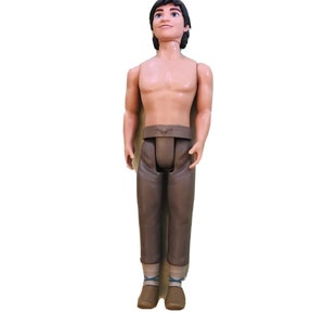 May include: A brown plastic doll with short black hair, wearing brown trousers and brown shoes. The doll has a sculpted face with brown eyes and a smile.