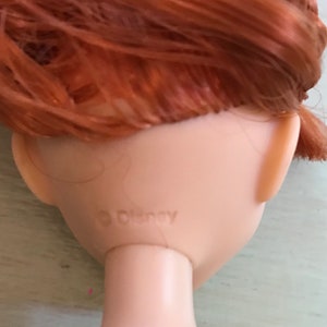 Puede incluir: Primer plano de la parte posterior de la cabeza de una mu&ntilde;eca con cabello rojo. La mu&ntilde;eca tiene un logotipo de Disney en la parte posterior del cuello.