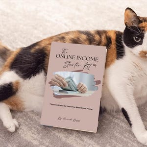 May include: A calico cat rests beside a book titled "The Online Income Starter Kit." The book cover is light pink with text and an image of hands holding money. The cat has a mix of black, orange, and white fur.