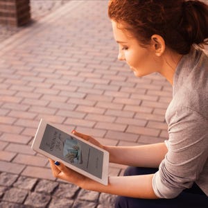 May include: A woman is holding a white tablet with a book cover visible on the screen. The book cover features text and an illustration. The woman is wearing a gray long-sleeved shirt and is sitting outside on a brick surface.