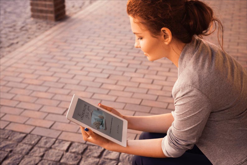 May include: A woman is holding a white tablet with a book cover visible on the screen. The book cover features text and an illustration. The woman is wearing a gray long-sleeved shirt and is sitting outside on a brick surface.