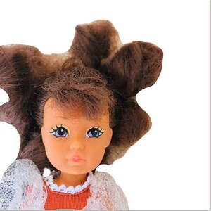 May include: A doll with brown hair, blue eyes, and a light brown complexion. She is wearing a white lace dress with a brown bodice.