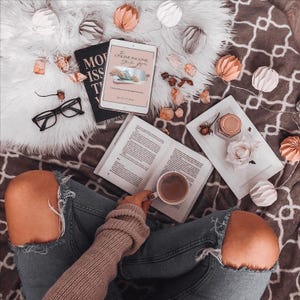May include: Overhead shot of a person in ripped jeans holding a coffee cup, with an open book, a tablet displaying "The Online Income Starter Kit", and a pair of black glasses. Decorative paper ornaments and a white tray with a rose and jar are also present.