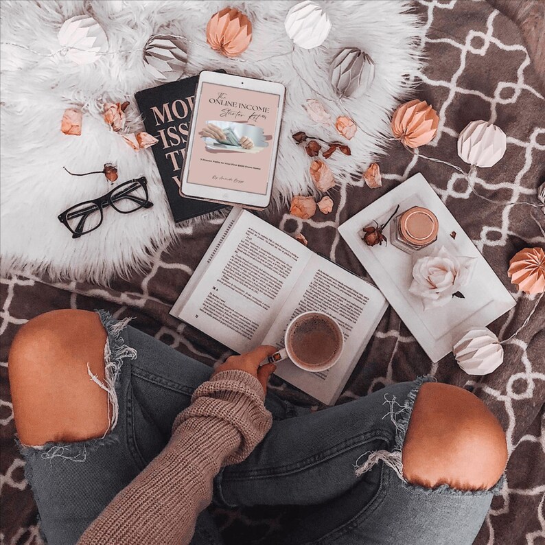 May include: Overhead shot of a person in ripped jeans holding a coffee cup, with an open book, a tablet displaying "The Online Income Starter Kit", and a pair of black glasses. Decorative paper ornaments and a white tray with a rose and jar are also present.