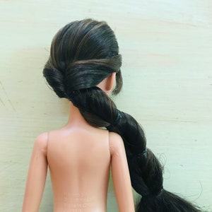 May include: A doll with long, dark brown hair styled in a braid. The doll is wearing no clothing and has a light skin tone. The doll's hair is tied with a black elastic band.
