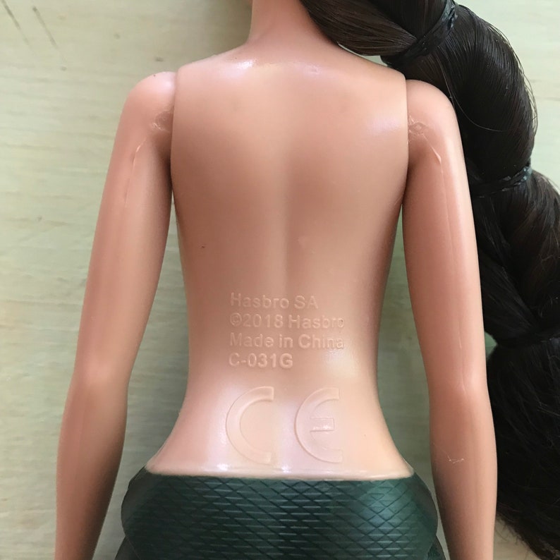 May include: A nude-toned doll with long dark brown hair. The doll is wearing a dark green skirt with a diamond pattern. The doll's back has the text "Hasbro SA &copy;2018 Hasbro Made in China C-031G" and a CE mark.