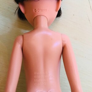 Op de afbeelding: Een naakte plastic pop met zwart haar, die een zwart topje draagt. De pop heeft een Disney-copyright op de achterkant van de nek en een Hasbro-label op de onderrug.
