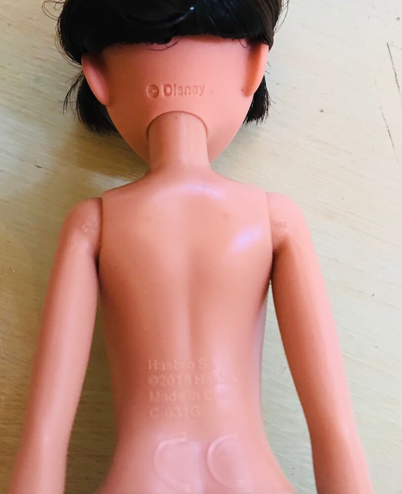 Op de afbeelding: Een naakte plastic pop met zwart haar, die een zwart topje draagt. De pop heeft een Disney-copyright op de achterkant van de nek en een Hasbro-label op de onderrug.