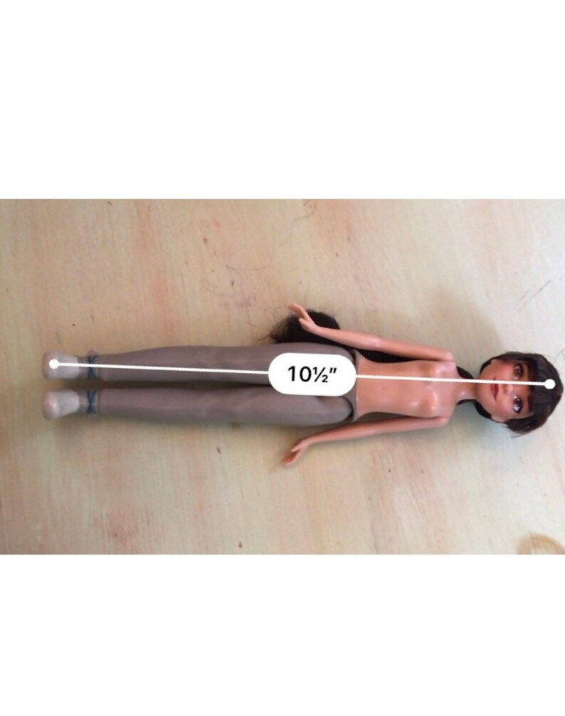 Op de afbeelding: Een pop met bruin haar en bruine ogen en een naakt bovenlichaam ligt op een lichtbruin oppervlak. De pop draagt een bruine broek en is 10 1/2 inch lang.