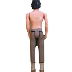 May include: A brown plastic action figure of a man with short black hair wearing brown trousers and brown boots. The figure has a barcode on its back.