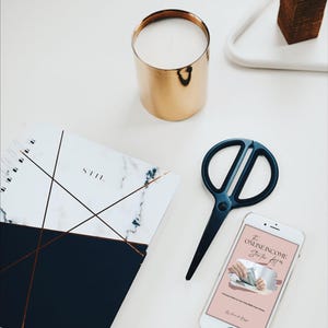 May include: A flat lay featuring a gold candle holder, a marble-patterned notebook, black scissors, and a smartphone displaying text. The notebook has a navy blue bottom half and copper-colored lines. The smartphone screen shows the text "The Online Income Starter Kit."