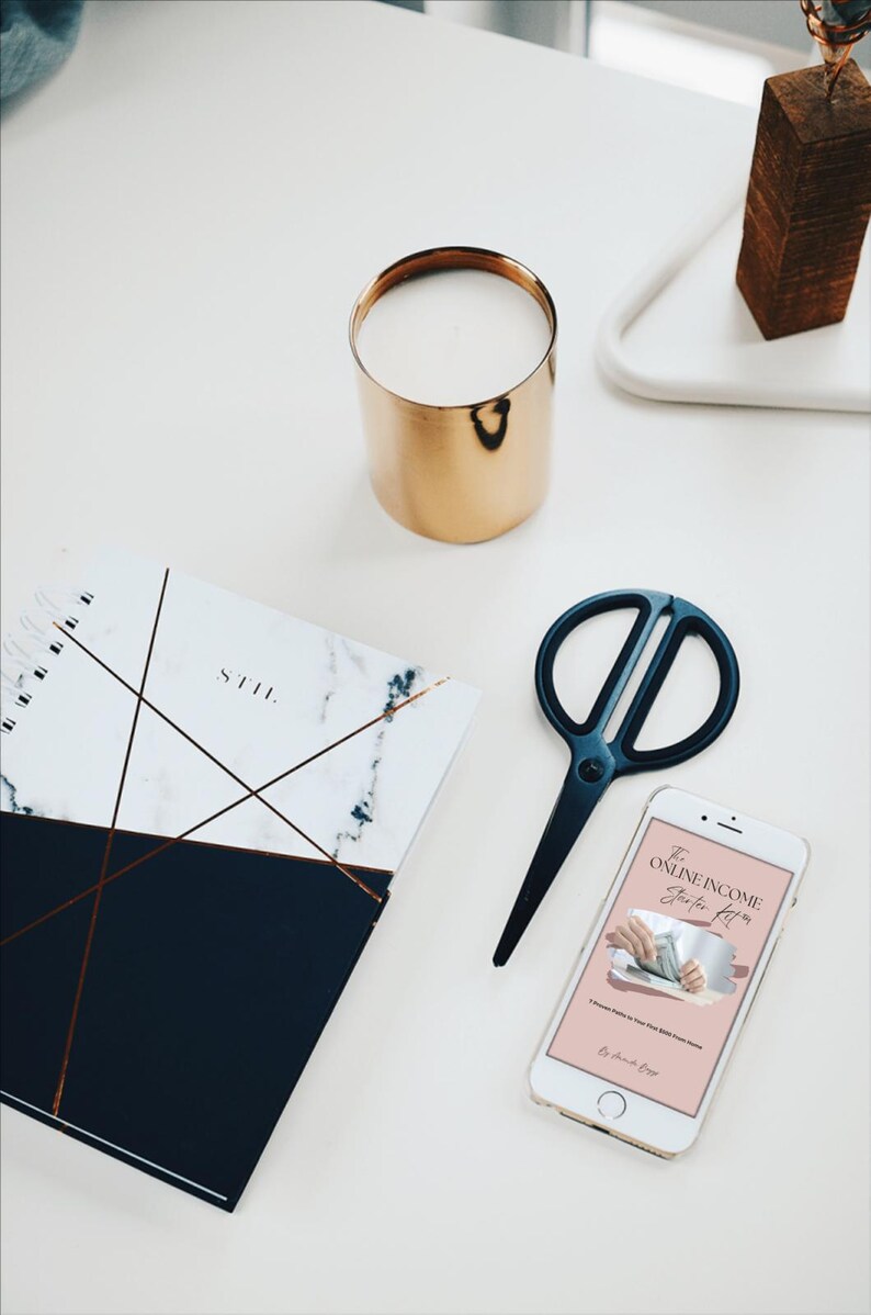 May include: A flat lay featuring a gold candle holder, a marble-patterned notebook, black scissors, and a smartphone displaying text. The notebook has a navy blue bottom half and copper-colored lines. The smartphone screen shows the text "The Online Income Starter Kit."