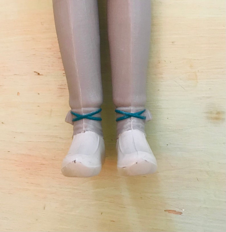 Op de afbeelding: Een paar witte poppen schoenen met turquoise blauwe veters. De schoenen zijn gemaakt van plastic en hebben een afgeronde neus.