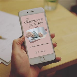 May include: A smartphone displays a pink screen with the text "The Online Income Starter Kit" and an image of hands holding money. The phone is held by a person, with a wooden table and papers in the background.