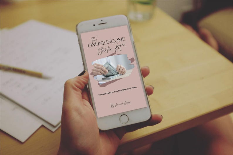May include: A smartphone displays a pink screen with the text "The Online Income Starter Kit" and an image of hands holding money. The phone is held by a person, with a wooden table and papers in the background.
