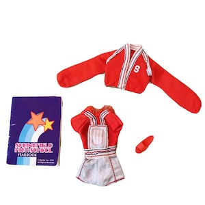 Puede incluir: Un conjunto de animadora rojo y blanco para una muñeca, que incluye un suéter rojo con una 'S' blanca en el frente, una falda roja y blanca con un borde blanco y azul, y un zapato rojo. El conjunto viene acompañado de un pequeño libro azul y morado con el texto 'Springfield High School Yearbook'.