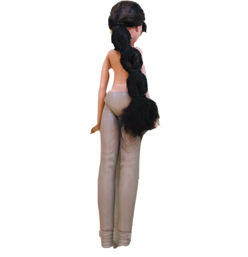 Op de afbeelding: Een pop met lang zwart haar in een vlecht, die een grijze broek en een grijs onderkleed draagt.