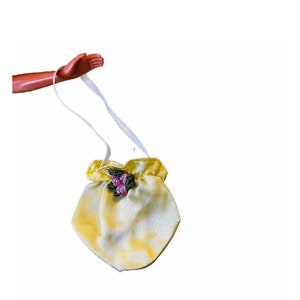 May include: A small, yellow drawstring bag with a white ribbon handle. The bag has a tie-dye pattern and is decorated with a cluster of pink and green floral embroidery. The bag is held by a brown arm.