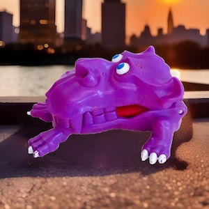 Peut inclure: Un monstre en plastique violet avec une grande bouche et des dents blanches. Le monstre a des yeux bleus et une langue rouge. Le jouet est posé sur une surface brune.