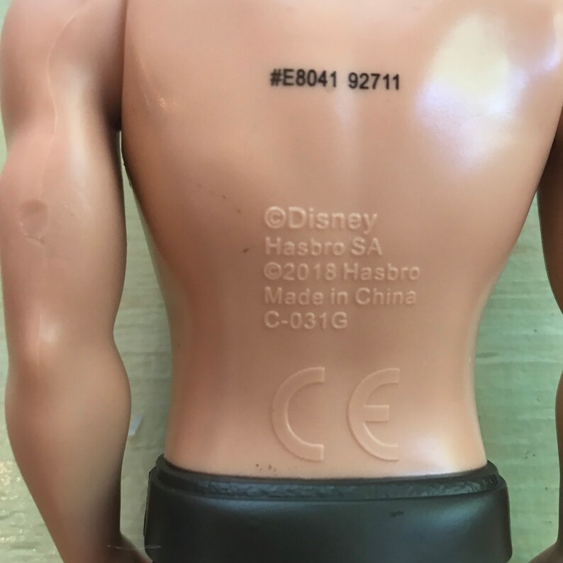 Puede incluir: Vista posterior de una figura de acci&oacute;n de pl&aacute;stico con un tono de piel bronceado. La figura lleva una prenda marr&oacute;n y negra. La figura tiene el siguiente texto impreso en la parte posterior: #E8041 92711 &copy;Disney Hasbro SA &copy;2018 Hasbro Hecho en China C-031G CE