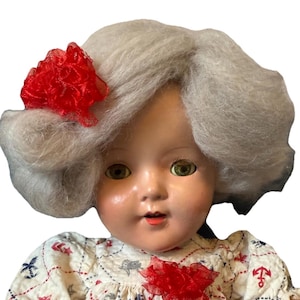 Op de afbeelding: Een vintage pop met een porseleinen gezicht, groene ogen en grijs haar gestyled in zachte golven. Een rode, gerimpelde stoffen bloem siert het haar. De pop draagt een witte jurk met een nautische print van ankers en zeilboten, en een rode stoffen bloem.