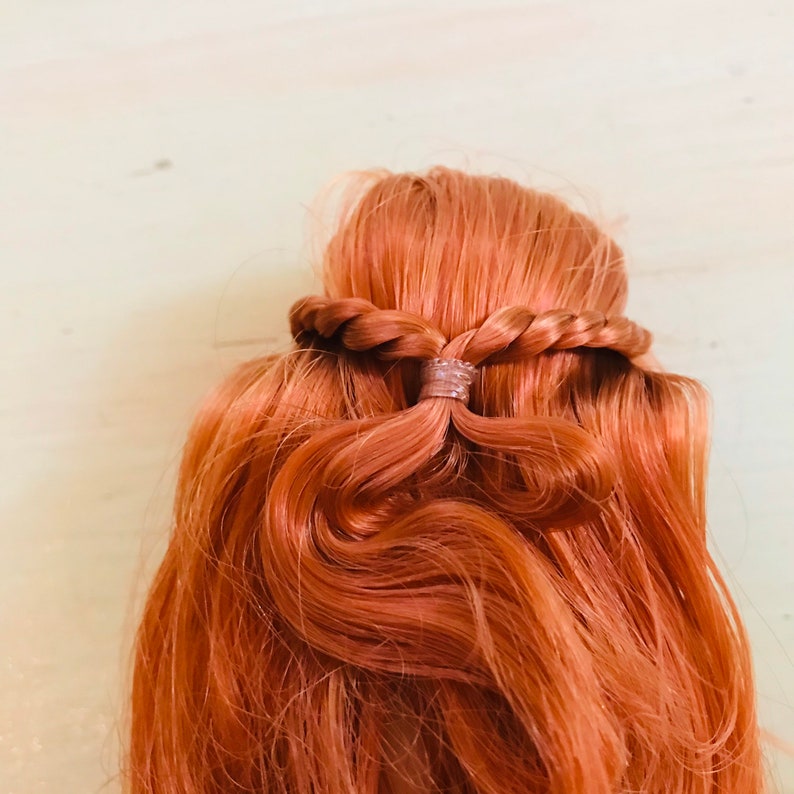 Puede incluir: Una mu&ntilde;eca con el pelo rojo peinado en una trenza, asegurada con una banda el&aacute;stica transparente.