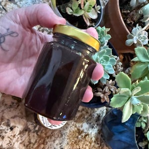 May include: A small, clear glass jar with a gold lid, filled with a dark brown liquid. The jar is held in a hand, with a blurred background of potted succulents. The jar appears to be sealed.