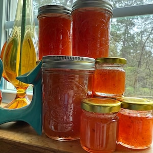 May include: Five glass jars filled with a red, translucent jam. The jars are arranged in a pyramid shape on a wooden surface. The jars have silver and gold lids.